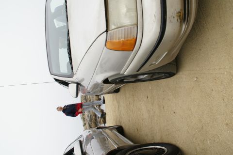 1990 Ford Taurus - A person stands between two cars, one a silver sedan, parked on a dirt surface.