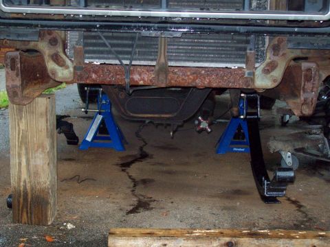1989 Ford F-150 - Front suspension of a truck is supported by jack stands, revealing rusty frame components and leaf springs.