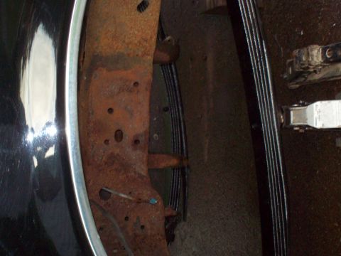 1989 Ford F-150 - This image shows the heavily rusted frame and leaf spring suspension of a truck, with a portion of a shiny fender visible.