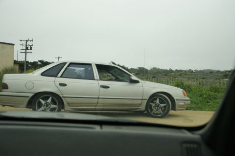 1990 Ford Taurus - Side view of a white sedan with aftermarket wheels on a dirt road.