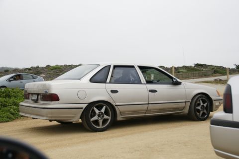 1990 Ford Taurus - A tan 1990 Ford Taurus sedan is parked on a dirt road, with another vehicle visible in the foreground and a light blue car in the backgroun…