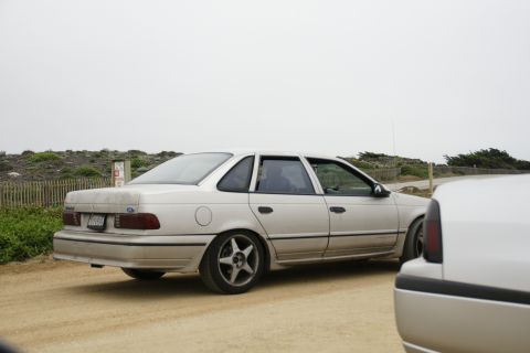 1990 Ford Taurus - Side view of a 1990 Ford Taurus sedan parked on a dirt road with another vehicle in the foreground.