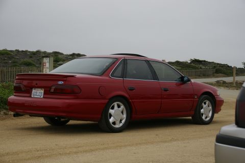 1990 Ford Taurus - A red 1990 Ford Taurus sedan is parked on a dirt road with a coastal background.