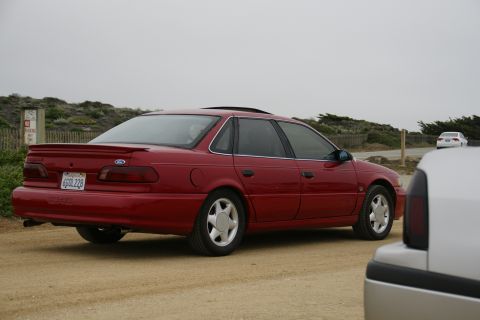 1990 Ford Taurus - Red 1990 Ford Taurus sedan parked on a dirt road with another car in the foreground and background.