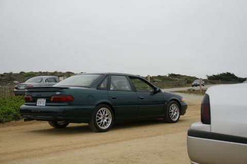 1990 Ford Taurus - A dark green sedan is parked on a dirt road with other vehicles nearby under an overcast sky.