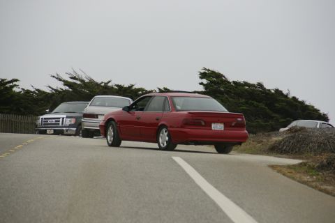 1990 Ford Taurus - A red 1990 Ford Taurus sedan is seen from the rear, driving on a road with a Ford pickup and another sedan nearby.