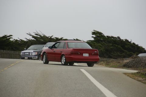 1990 Ford Taurus - A red 1990 Ford Taurus sedan is seen from the rear, with a Ford pickup truck behind it on the road.