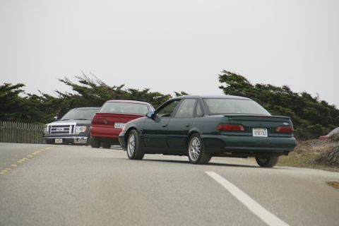 1990 Ford Taurus - Three vehicles, including a 1990 Ford Taurus, drive on a road with trees and a fence.