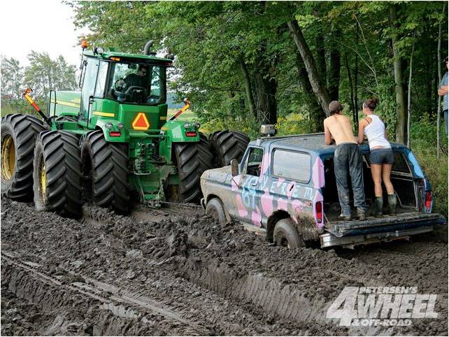 Blue Ford Bronco with pink and black camouflage, stuck in mud, being pulled by a green tractor