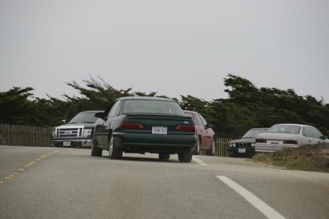 1990 Ford Taurus - A dark green 1990 Ford Taurus sedan is seen from the rear on a road, with other vehicles and trees in the background.