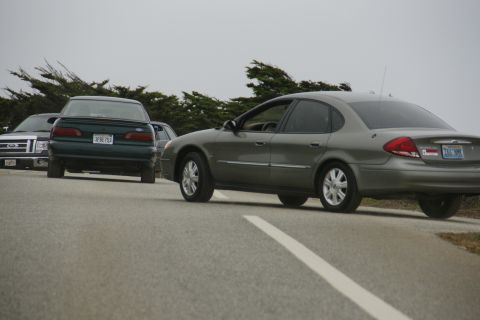 1990 Ford Taurus - Multiple vehicles are parked or driving on a road with trees in the background under an overcast sky.