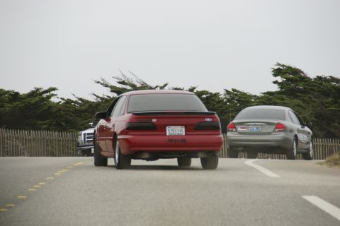 1990 Ford Taurus - Two sedans drive down a road with a wooden fence and trees lining the side under an overcast sky.
