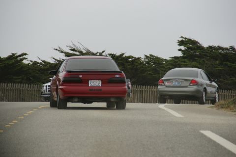 1990 Ford Taurus - Two sedans drive on a road with a wooden fence and trees in the background.
