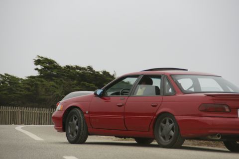 1990 Ford Taurus - A red 1990 Ford Taurus sedan is seen from the side on a road with trees in the background.