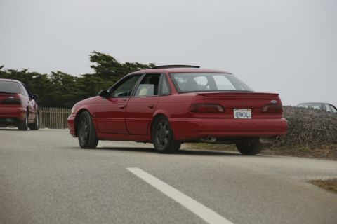 1990 Ford Taurus - A red sedan is parked on the side of the road, with another red car visible to its left.
