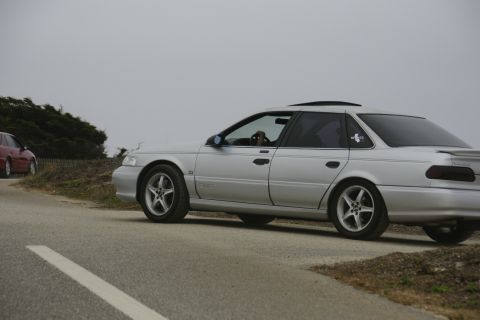 1990 Ford Taurus - A silver 1990 Ford Taurus sedan is parked on the side of a road with another car in the background.