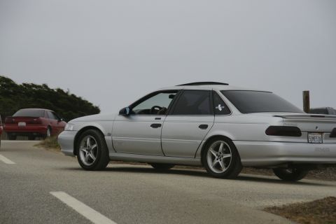 1990 Ford Taurus - A silver 1990 Ford Taurus sedan is seen from the side, with another red car visible in the background on a cloudy day.