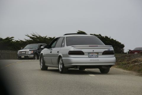 1990 Ford Taurus - A silver sedan drives down a road with a pickup truck in the background under an overcast sky.