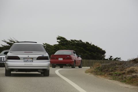 1990 Ford Taurus - A silver 1990 Ford Taurus is followed by a red sedan on a coastal road under an overcast sky.