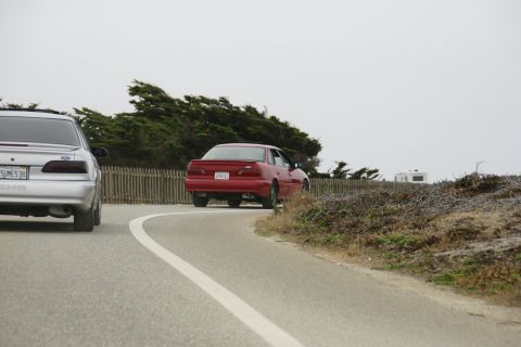 1990 Ford Taurus - A red sedan drives away on a winding road with a silver car in the foreground and a trailer in the background.