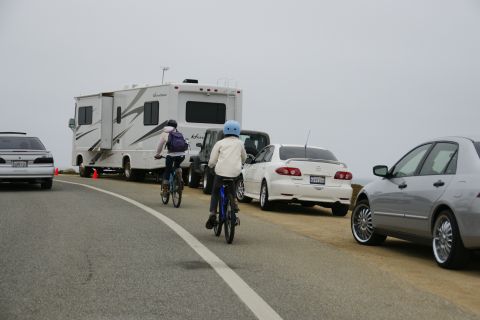1990 Ford Taurus - A silver sedan is parked on the side of the road next to other vehicles and people on bicycles.