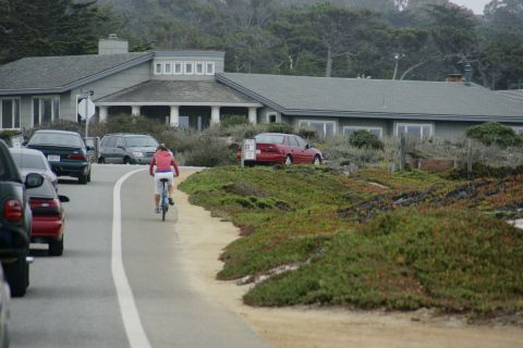 1990 Ford Taurus - A red sedan is parked on the side of the road near a coastal house, with a cyclist passing by.