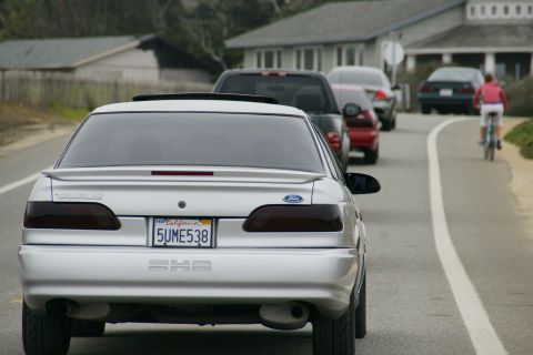 1990 Ford Taurus - A silver 1990 Ford Taurus SHO is seen from behind in traffic, with other vehicles and a cyclist nearby.