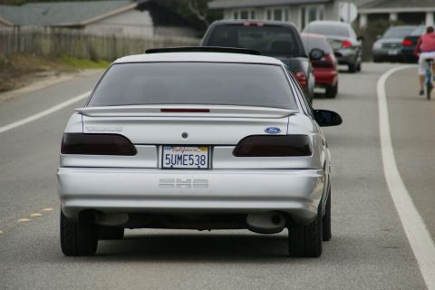 1990 Ford Taurus - A silver 1990 Ford Taurus drives down a road, with other vehicles and a cyclist visible.