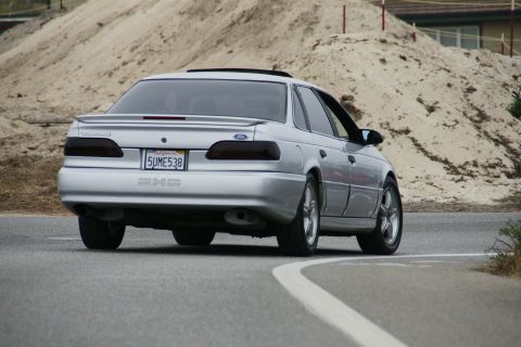 1990 Ford Taurus - This silver sedan is seen from the rear on a winding road with a sandy embankment in the background.