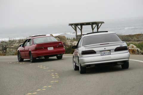 1990 Ford Taurus - Two Ford Taurus sedans drive along a scenic coastal highway under an overcast sky.