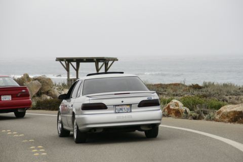1990 Ford Taurus - A silver 1990 Ford Taurus drives along a coastal road, with another red car visible behind it.