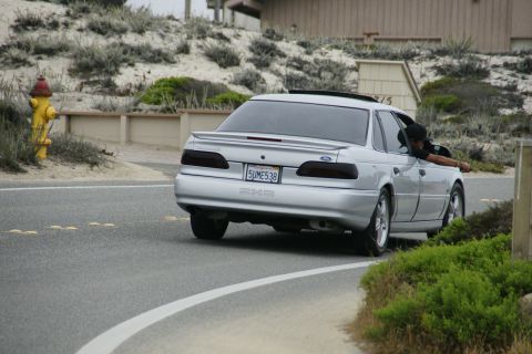 1990 Ford Taurus - A silver sedan drives down a coastal road with sand dunes and vegetation on the side.