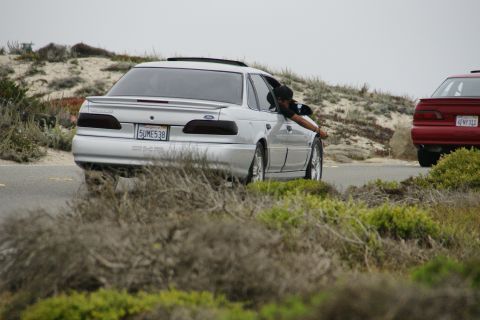 1990 Ford Taurus - A silver 1990 Ford Taurus sedan is seen from the rear, with a person leaning out the driver's side window as it drives on a coastal road.