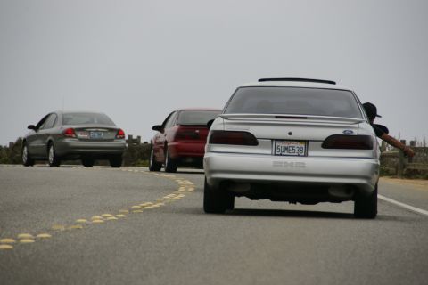 1990 Ford Taurus - A silver car, a red car, and a gray car drive down a road with a wooden fence and overcast skies.