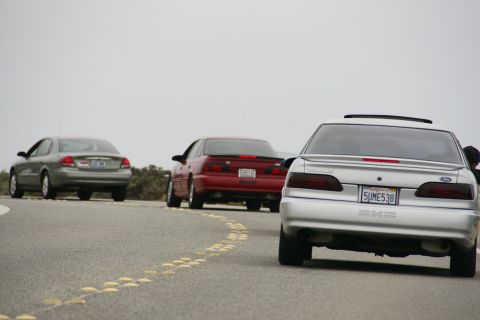 1990 Ford Taurus - Three sedans, including a silver 1990 Ford Taurus SHO, drive along a coastal road under an overcast sky.