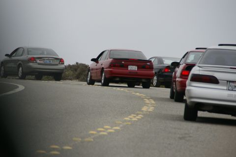 1990 Ford Taurus - A red 1990 Ford Taurus with a spoiler is seen from the rear, driving in a line with other vehicles.