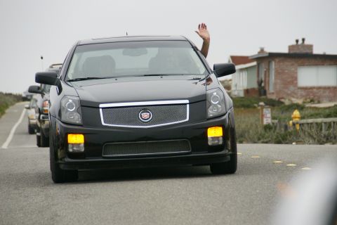 1990 Ford Taurus - Cadillac sedan driving on a coastal road with a person waving from the driver's side window.