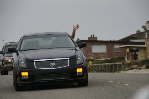 1990 Ford Taurus - A black Cadillac sedan is seen driving down a road with a person's hand waving from the window.