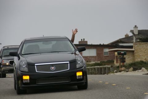 1990 Ford Taurus - A black Cadillac sedan drives down a coastal road, with a second vehicle visible behind it.