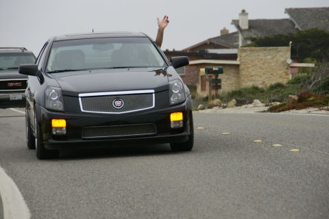 1990 Ford Taurus - A black Cadillac sedan drives down a coastal road, with a GMC truck visible in the background.
