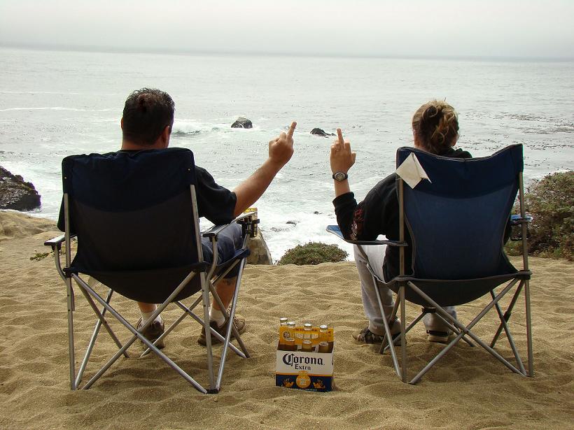 Two people sit in camping chairs on a sandy beach, facing the ocean and giving the middle finger. A six-pack of Corona Extra is on the sand between them