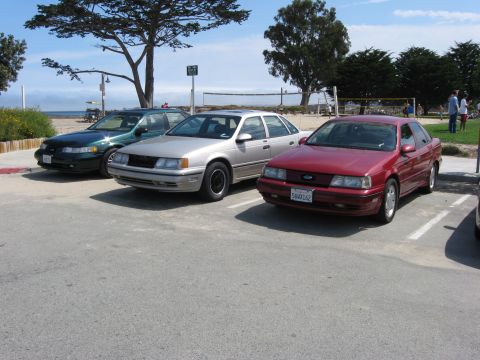 2003 Ford Taurus - Three Ford Taurus sedans are parked in a lot near a beach, with the red car in the foreground.