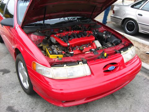 2003 Ford Taurus - The front of a red Ford sedan with its hood open, revealing the engine bay. A second car is visible in the background.