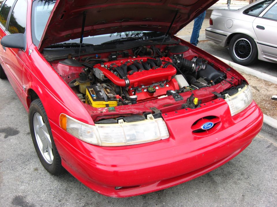 Red Ford sedan, hood open showing engine. Another car is parked behind it