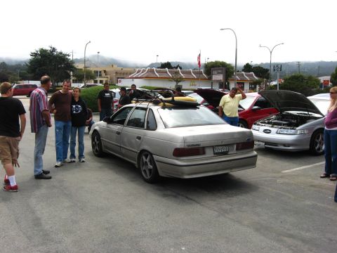 2003 Ford Taurus - A silver sedan with a roof rack is parked in a lot with other vehicles and people.