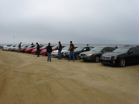 2003 Ford Taurus - A group of cars are parked on the beach near the water under an overcast sky.