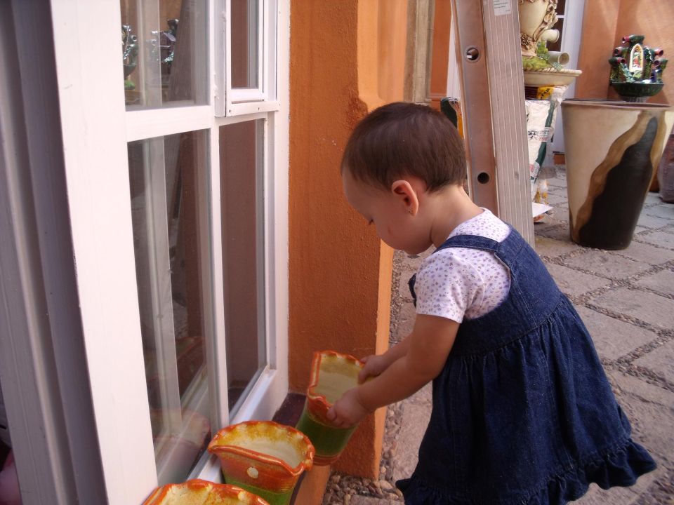 Child in denim overalls watering potted plants near a window