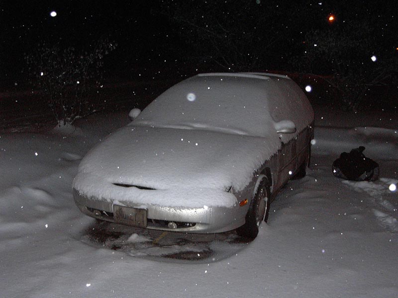 Silver Ford Taurus sedan covered in snow at night while snow falls