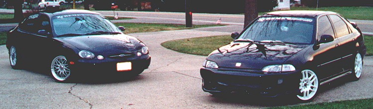 Two lowered dark sedans with windshield banners and aftermarket wheels in a driveway