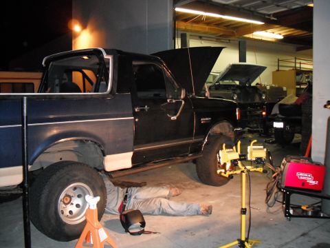 1991 Ford Bronco - A mechanic works on the underside of a 1991 Ford Bronco in a garage, with another vehicle visible in the background.